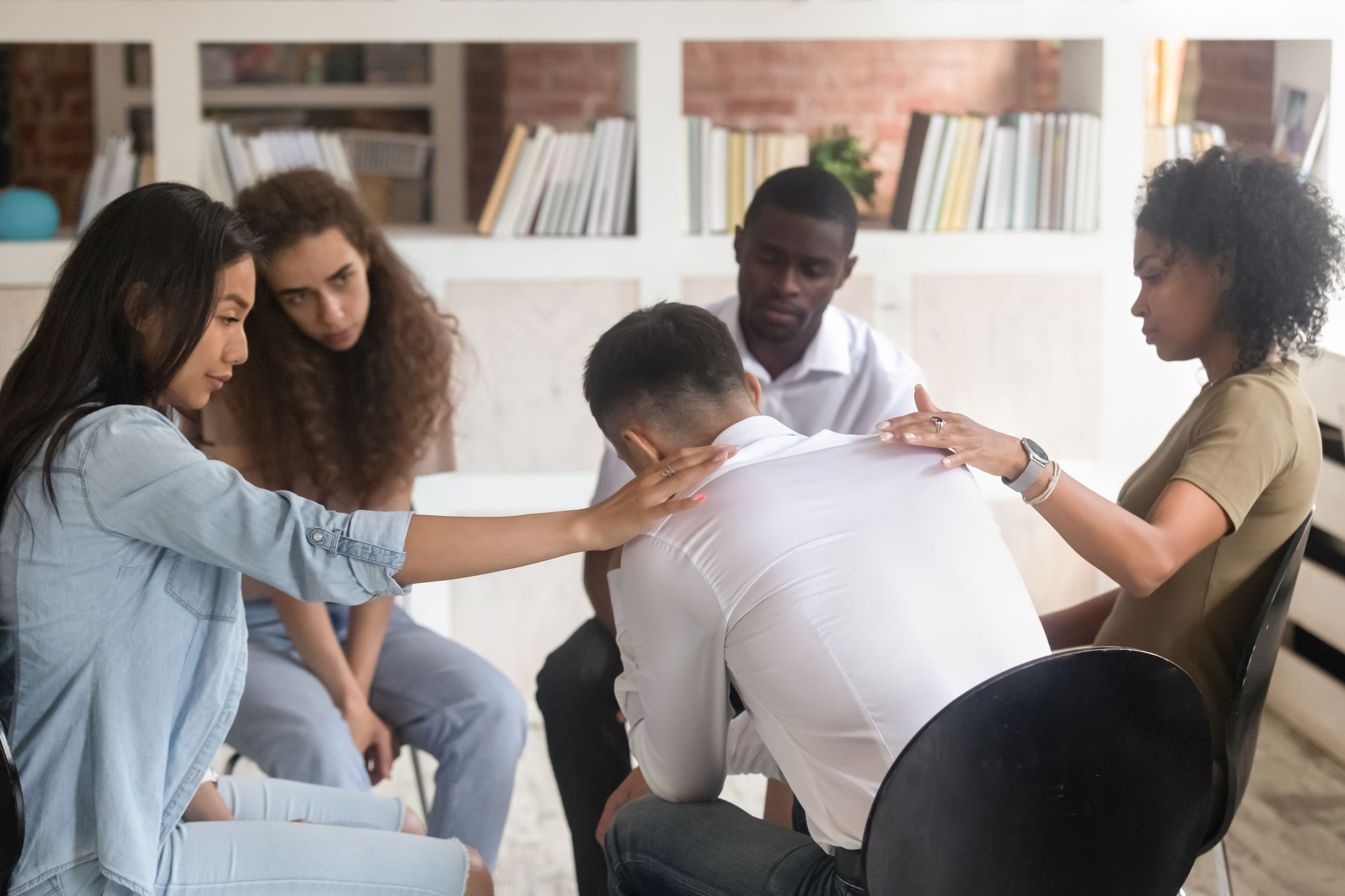 Back view of diverse people sit in circle at psychological therapy session, friends hug support show understanding to depressed man suffer from addiction have life trauma at psychotherapy counselling