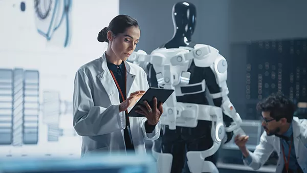 Two scientists in the workplace. One person is using a tablet while a second person checks a robotic arm.