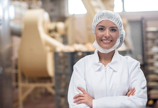 woman wearing white jacket, head covering and mask in an industrial food prep setting.