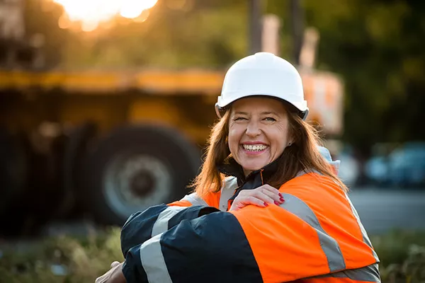 Woman wearing hat hat and orange jacket working outdoors at an industrial construction site.