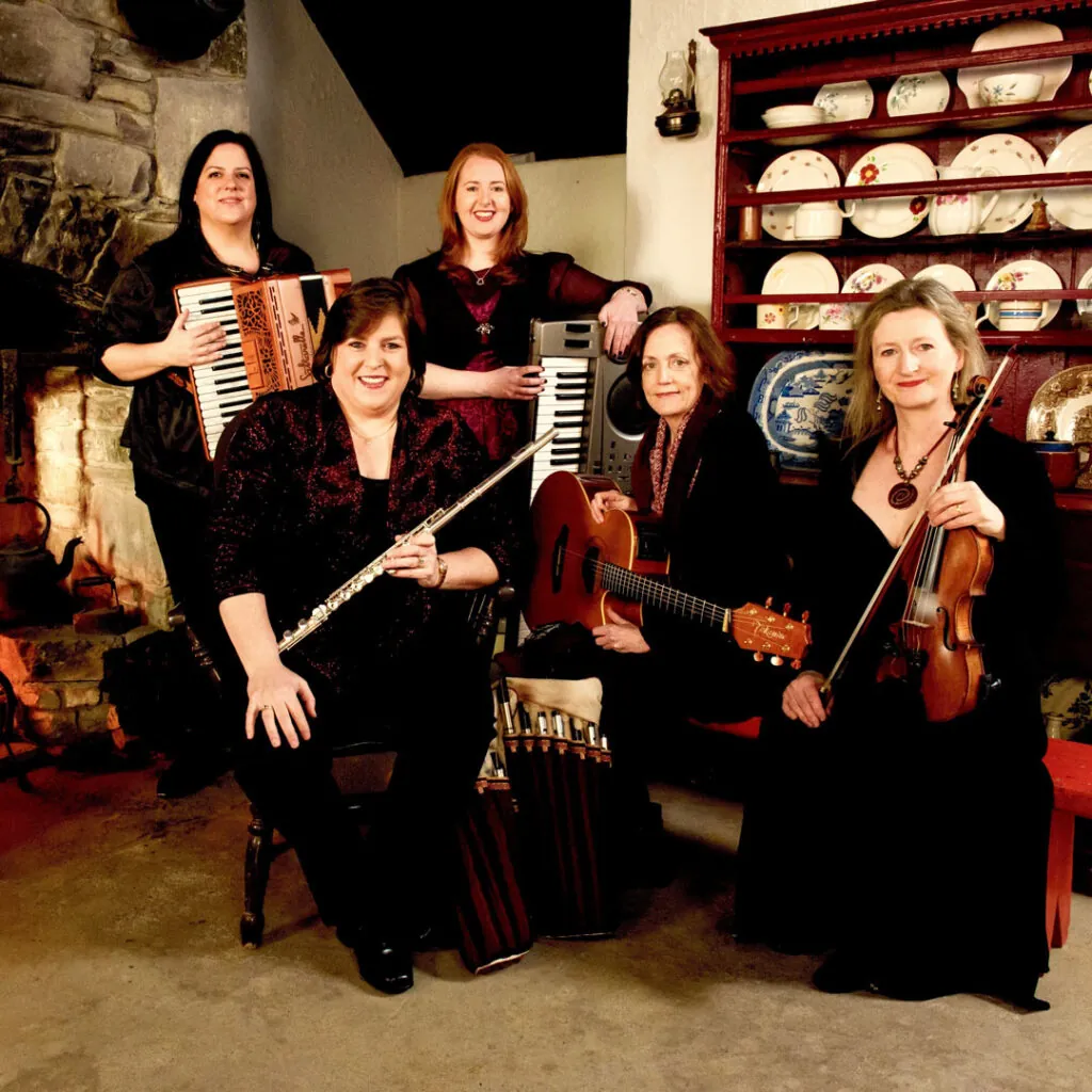 A group of five women with musical instruments in a cozy room, featuring an accordion, flute, keyboard, guitar, and violin.