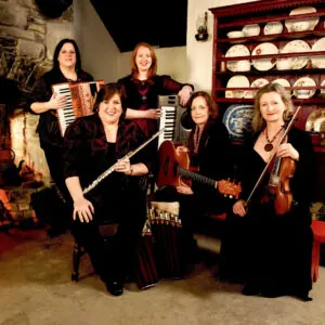 A group of five women with musical instruments in a cozy room, featuring an accordion, flute, keyboard, guitar, and violin.