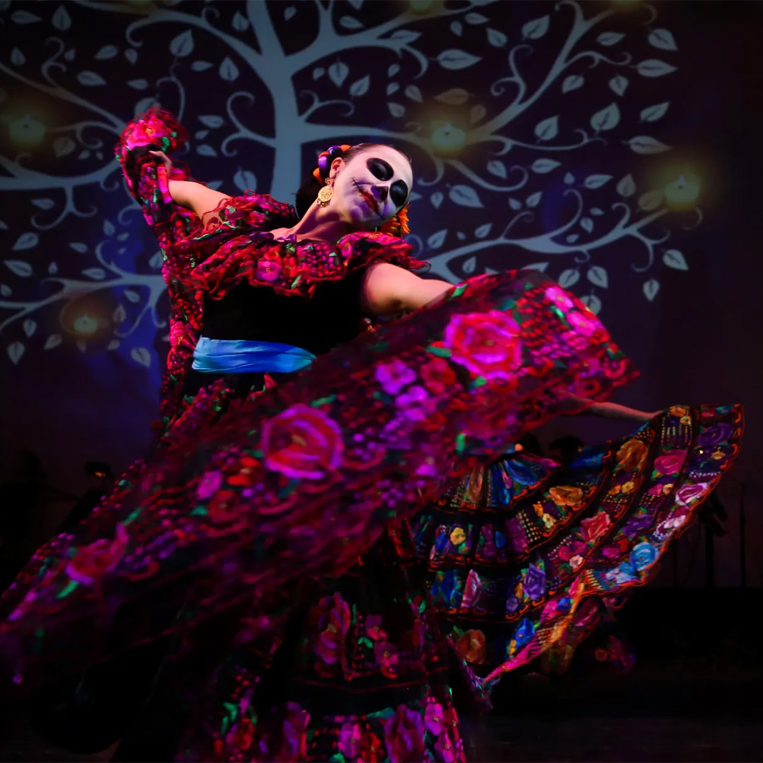 A dancer in colorful traditional dress and face paint performs on stage against a backdrop of a stylized tree.