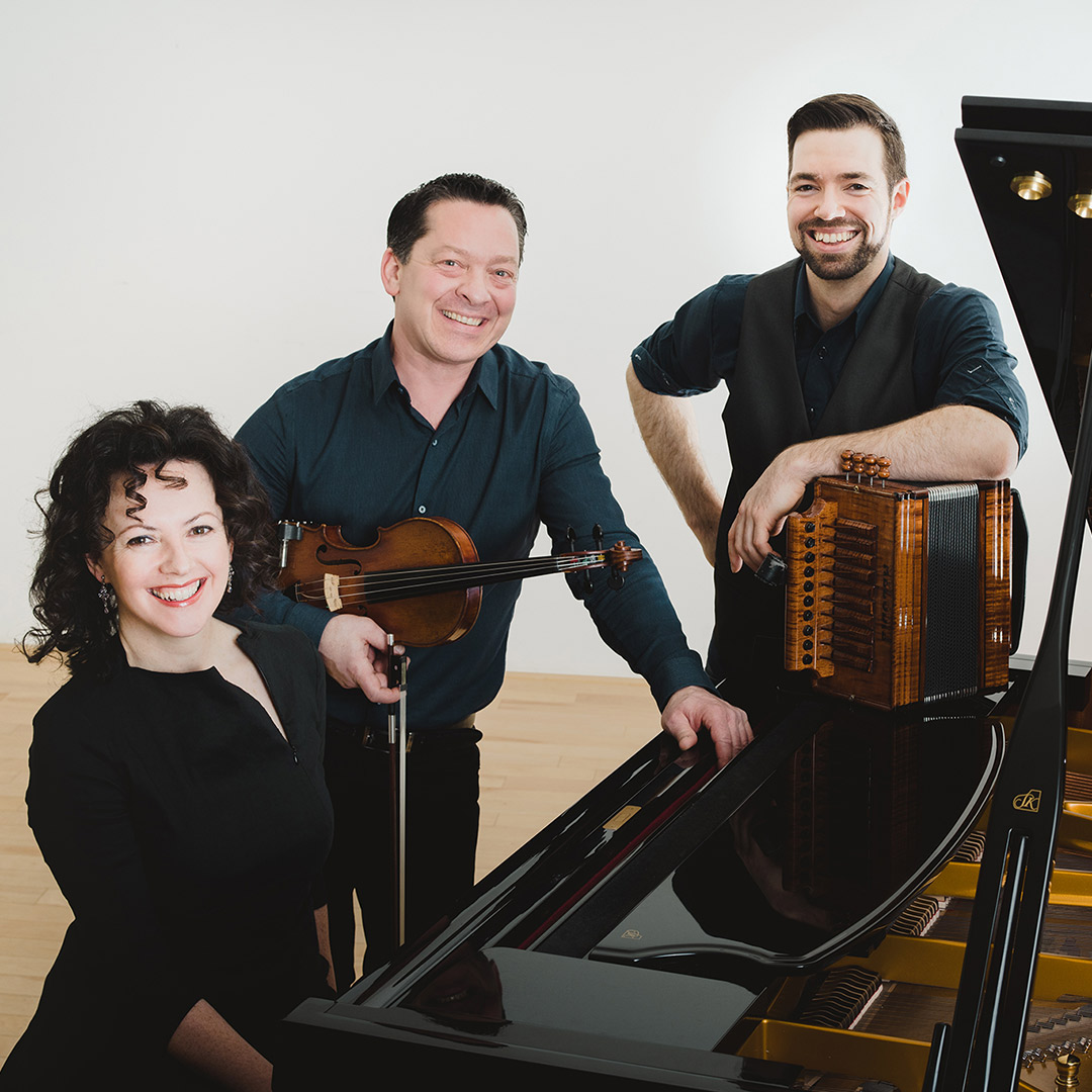 Three musicians smiling next to a grand piano, one holds a violin, and another has an accordion.