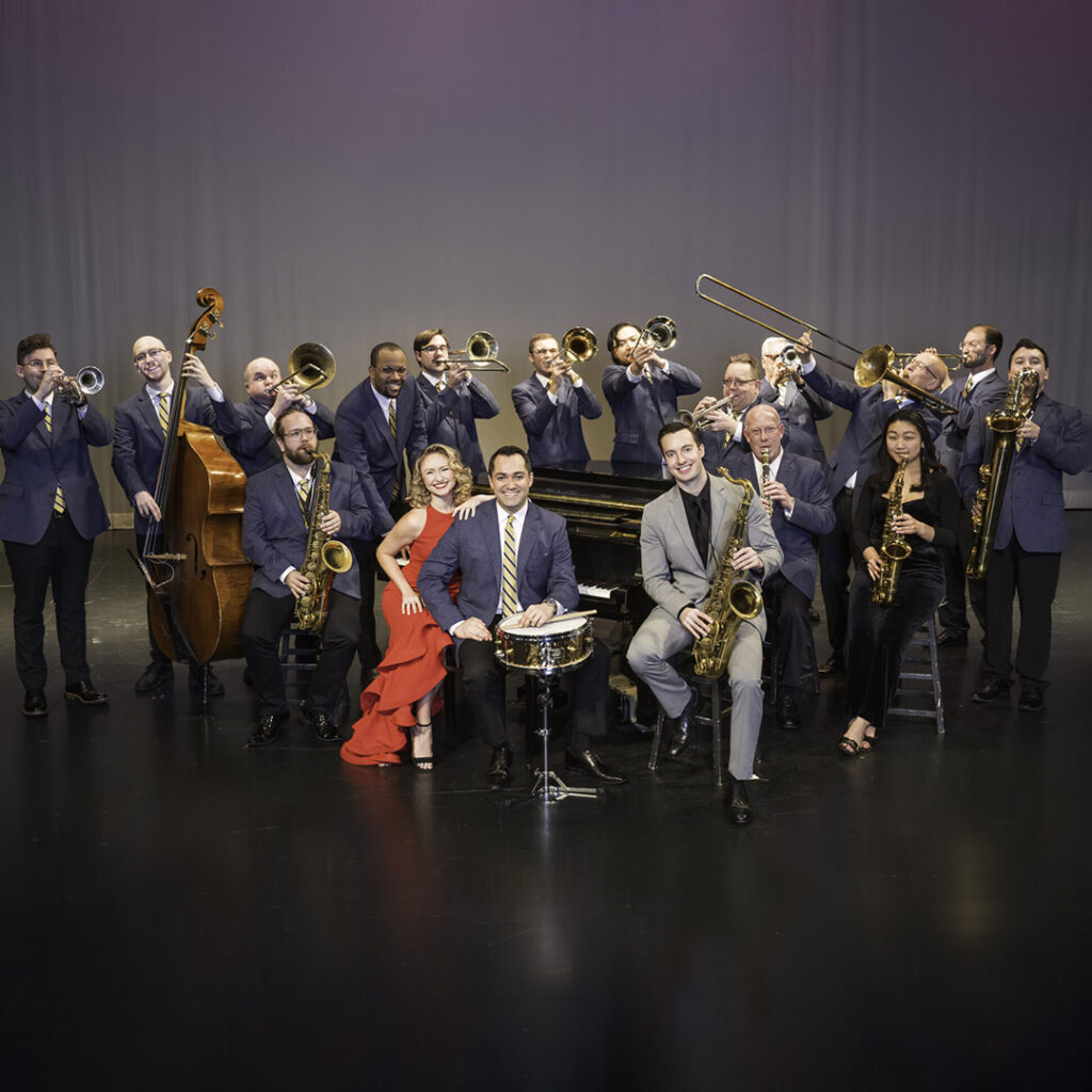 A jazz band with fifteen musicians on stage, wearing formal attire with various instruments including trumpets, trombones, saxophones, and a double bass.