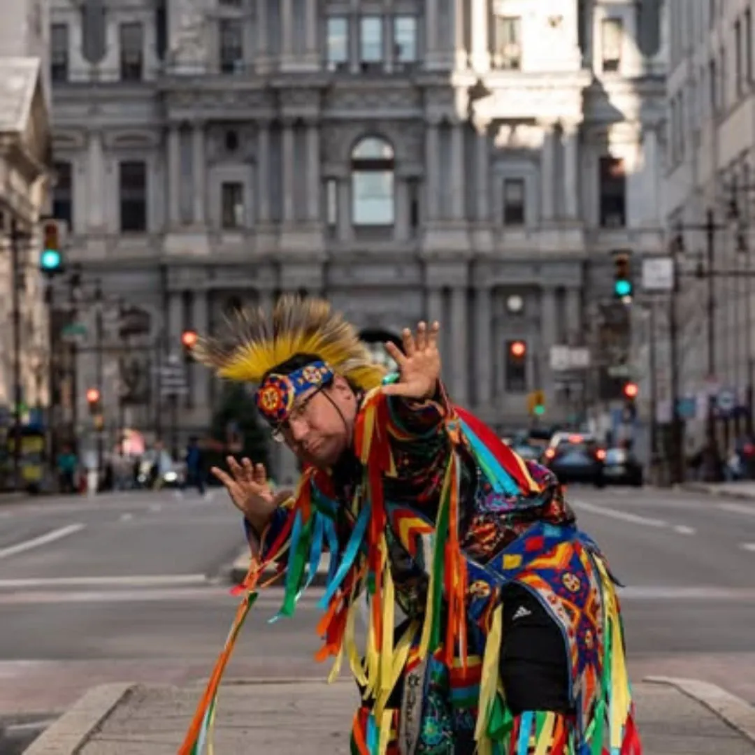 A person in colorful traditional regalia with a feather headdress stands on a city street with a large ornate building in the background.