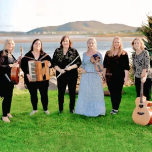 Six women stand on a green lawn, holding various musical instruments, with a scenic water and hill view in the background.