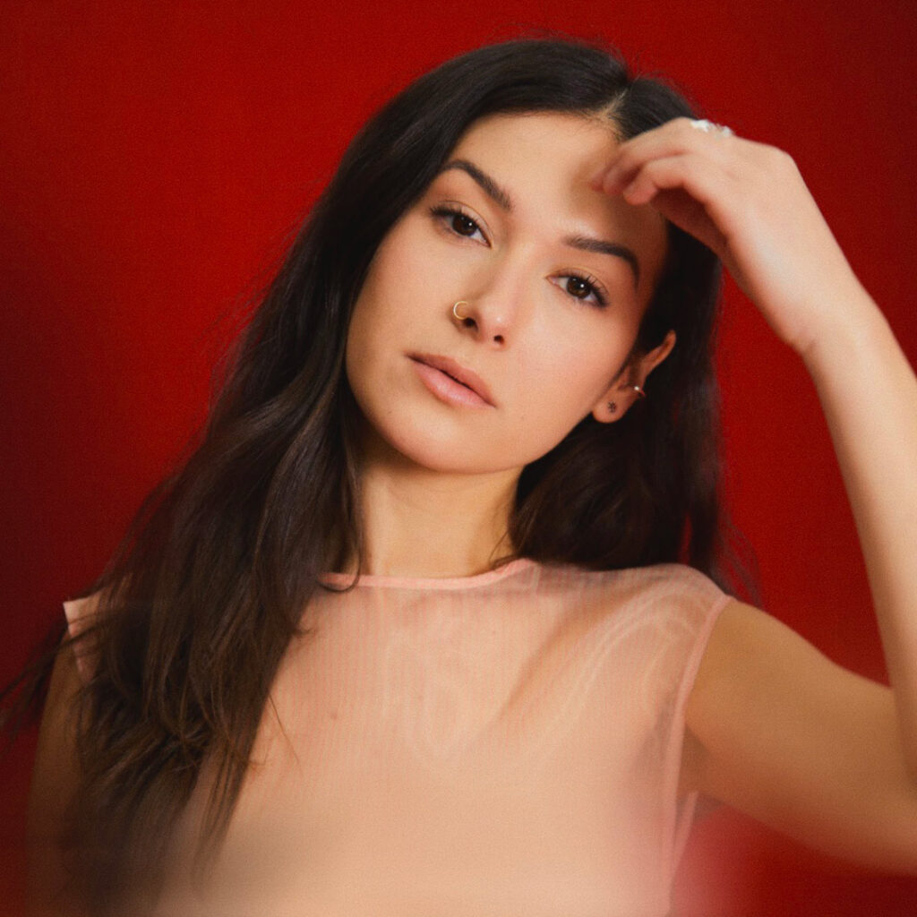 A person with long dark brown hair in a beige top in front of a red background