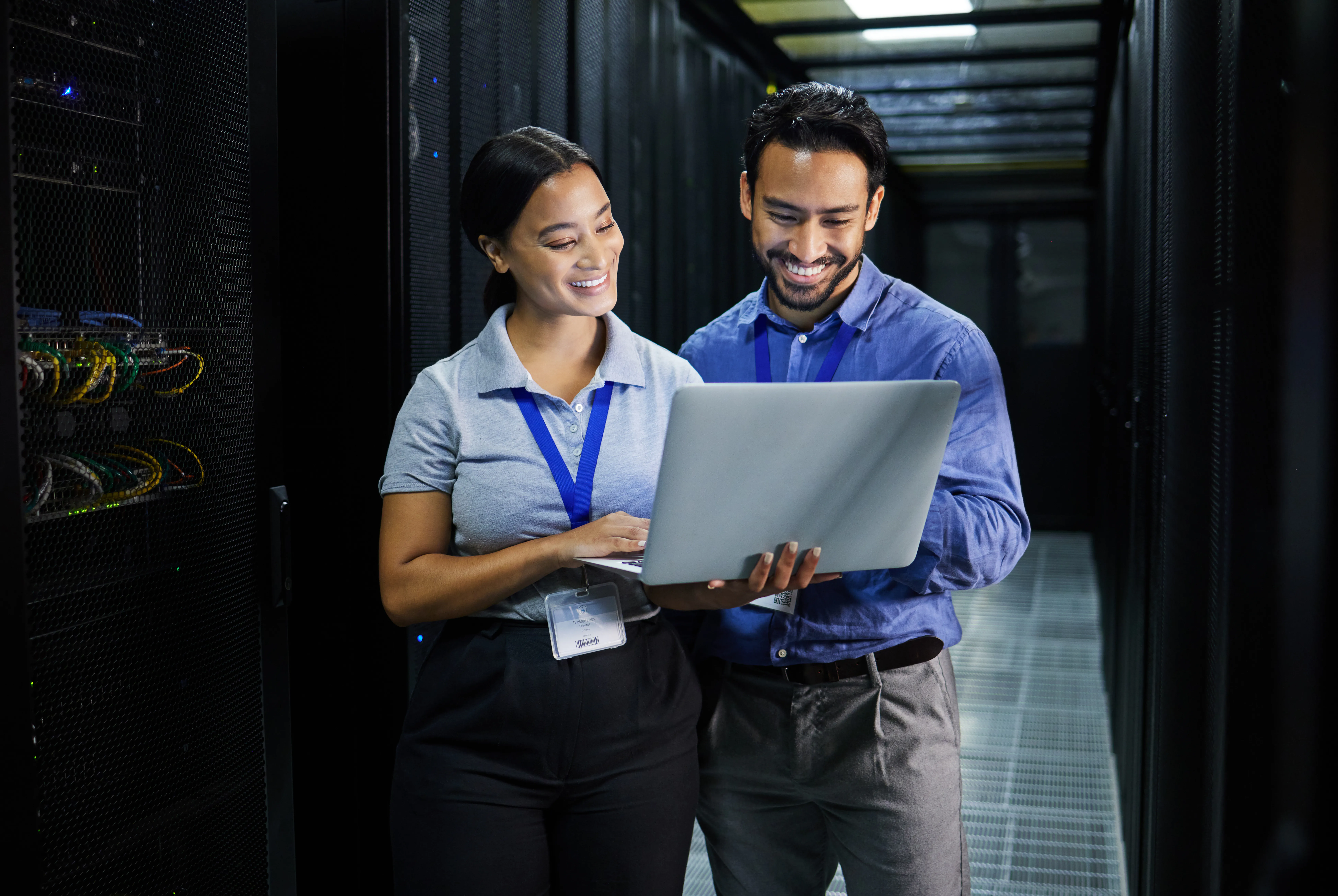 Two smiling IT professionals standing in a server room looking at a laptop