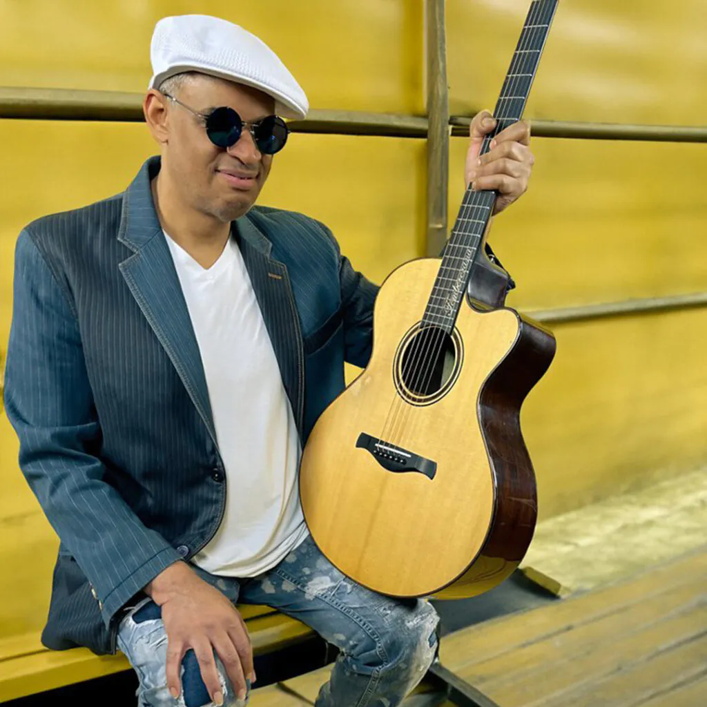 A person in a white hat and sunglasses holding a guitar in front of a yellow wall