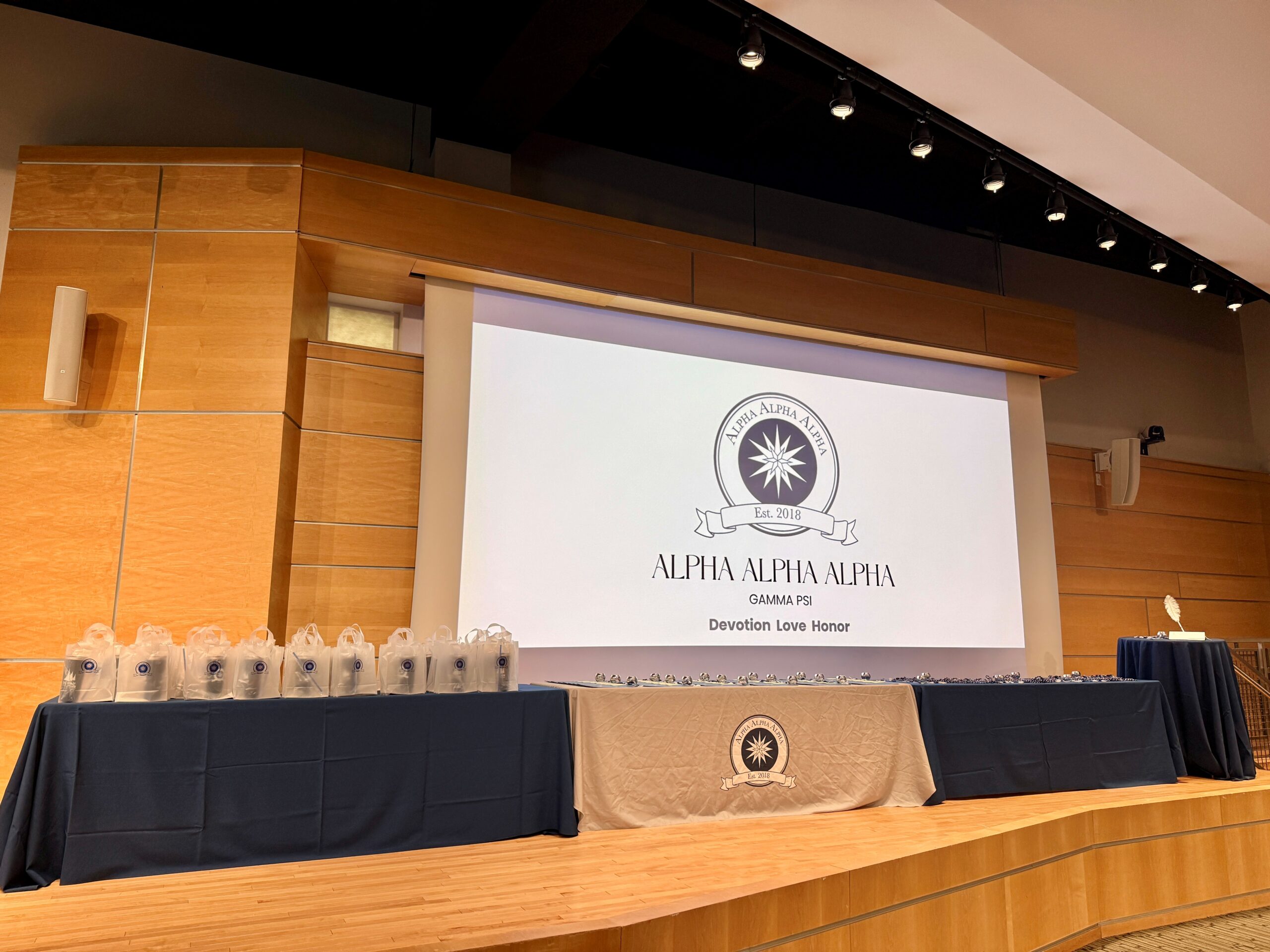 Stage with a projector screen displaying the Tri-Alpha logo and 4 tables laid with induction items