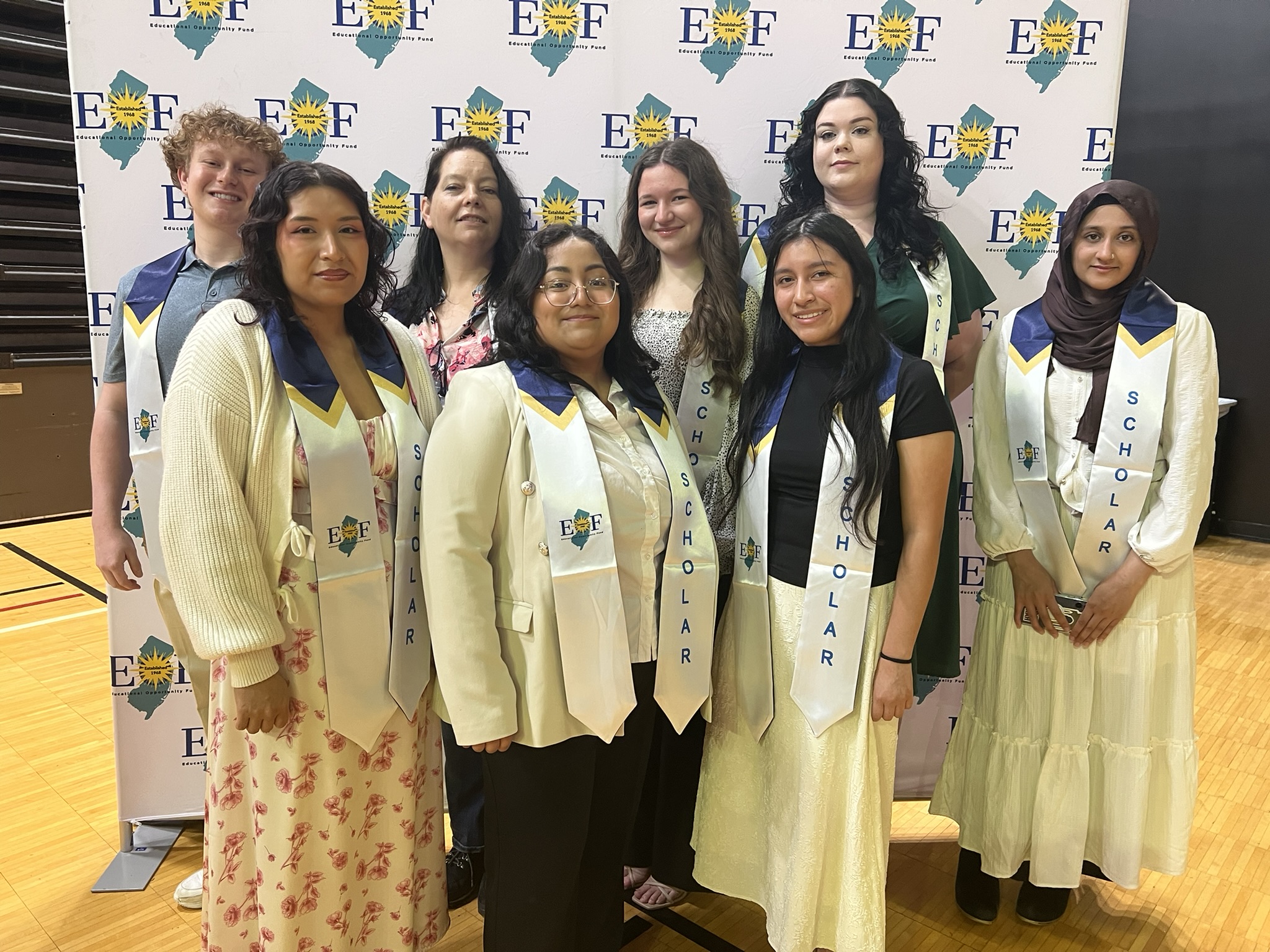 Group of EOF students in their white achievement stoles posing in front of an EOF step and repeat banner.