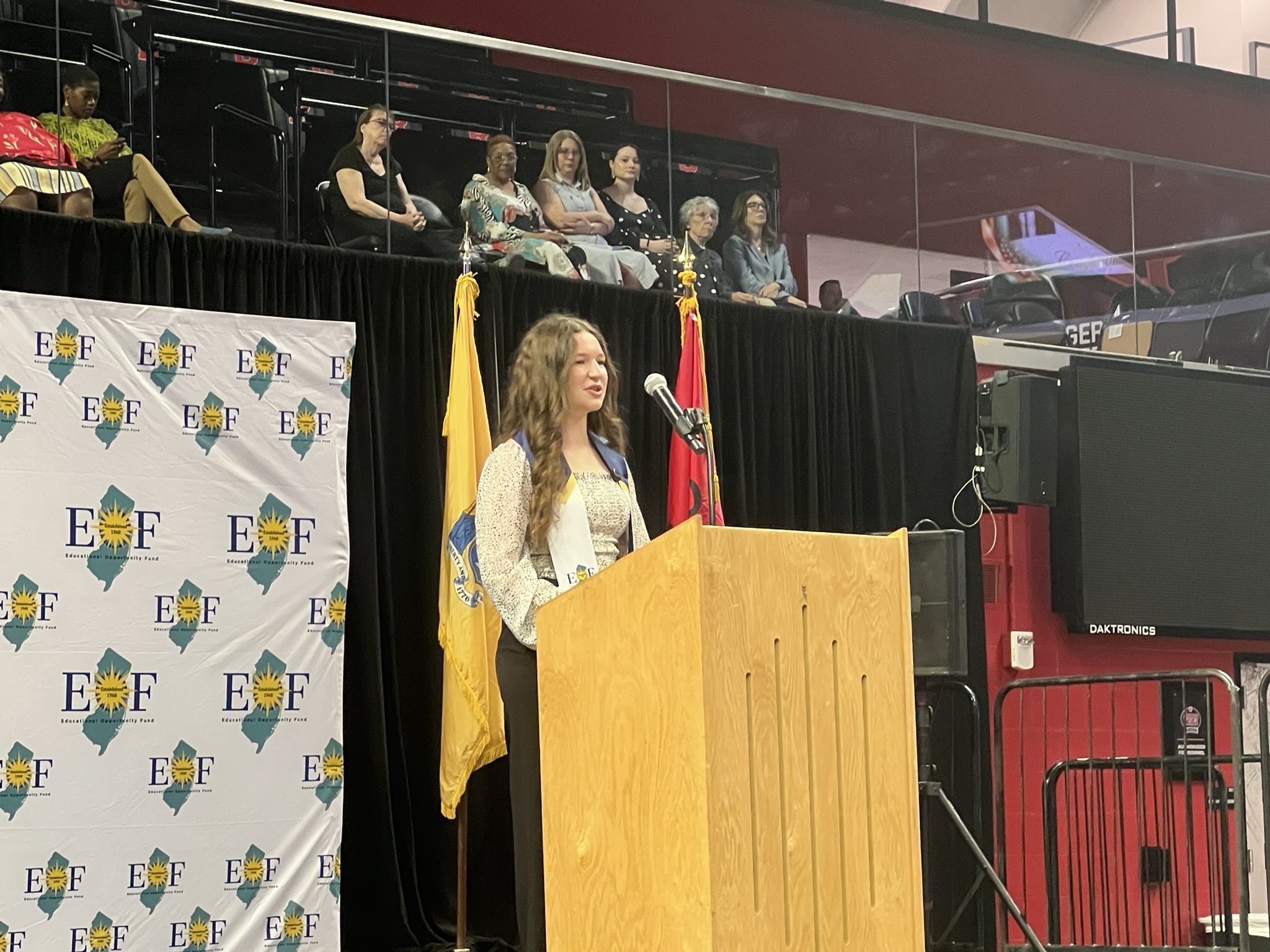 Female student with long brown hair stands at a wood podium giving a speech.