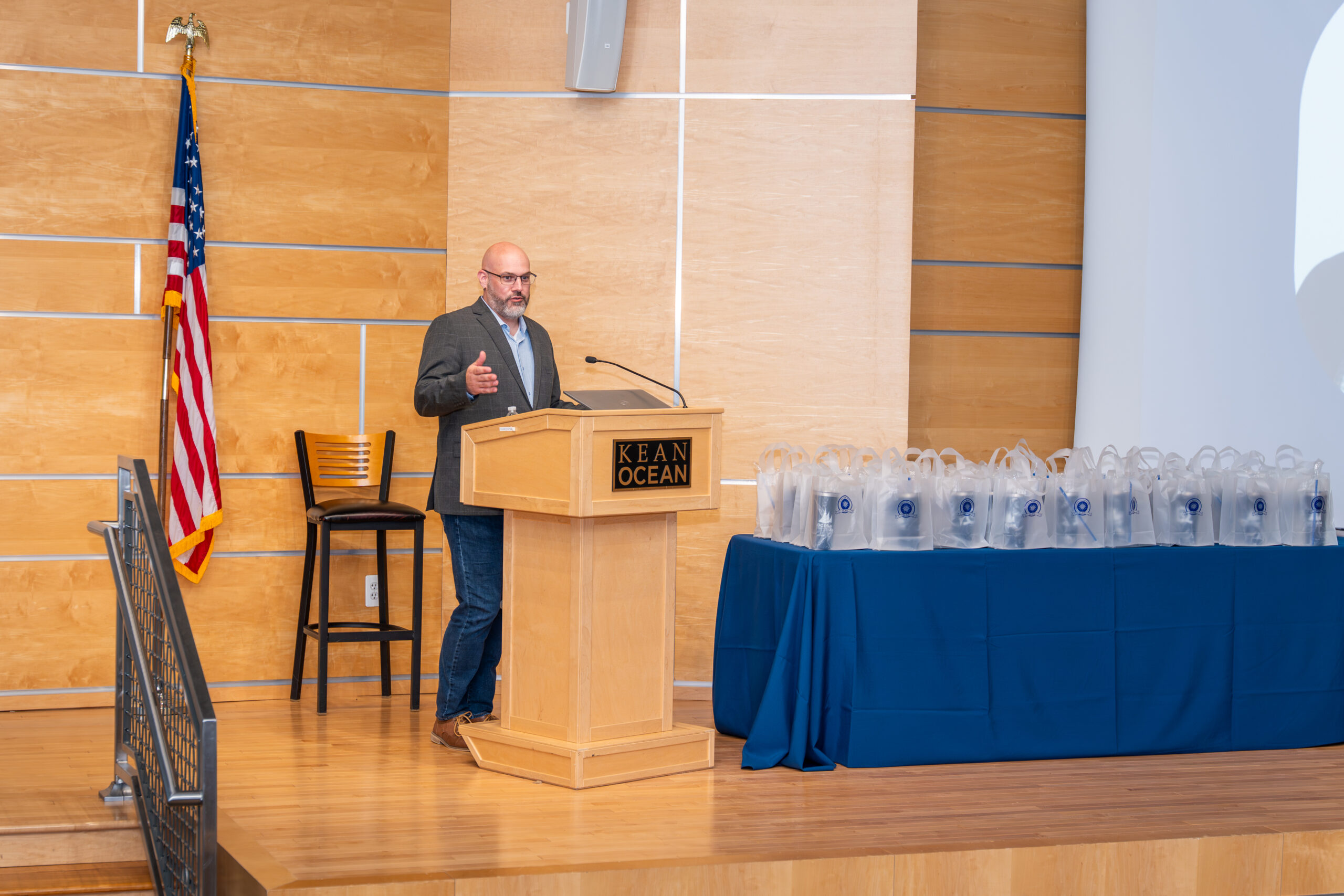 Man with glasses in grey blazer standing behind a podium giving a speech.