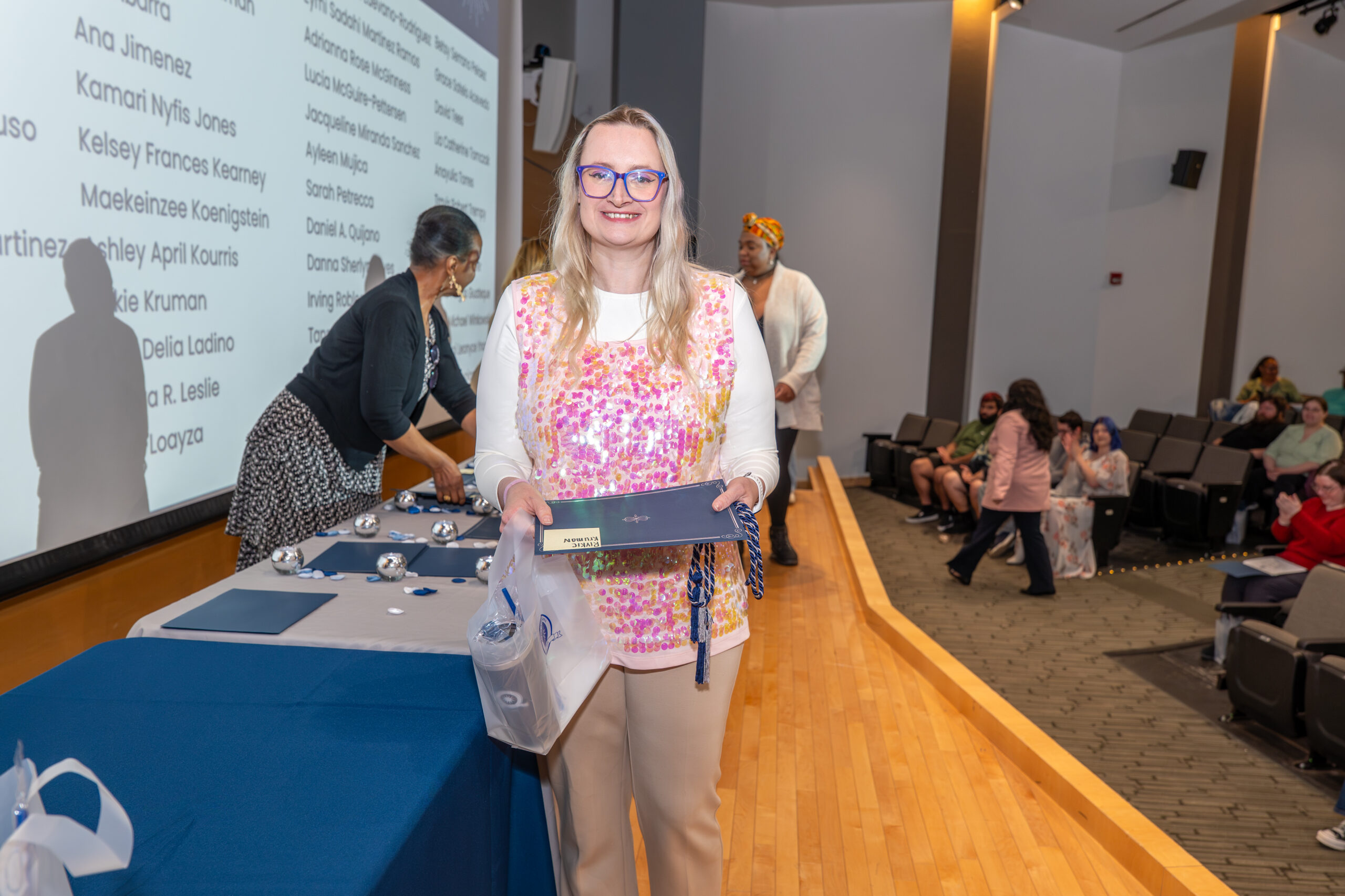 Student with blonde hair and glasses walking across a stage holding a certificate, honor cords, and a bag.