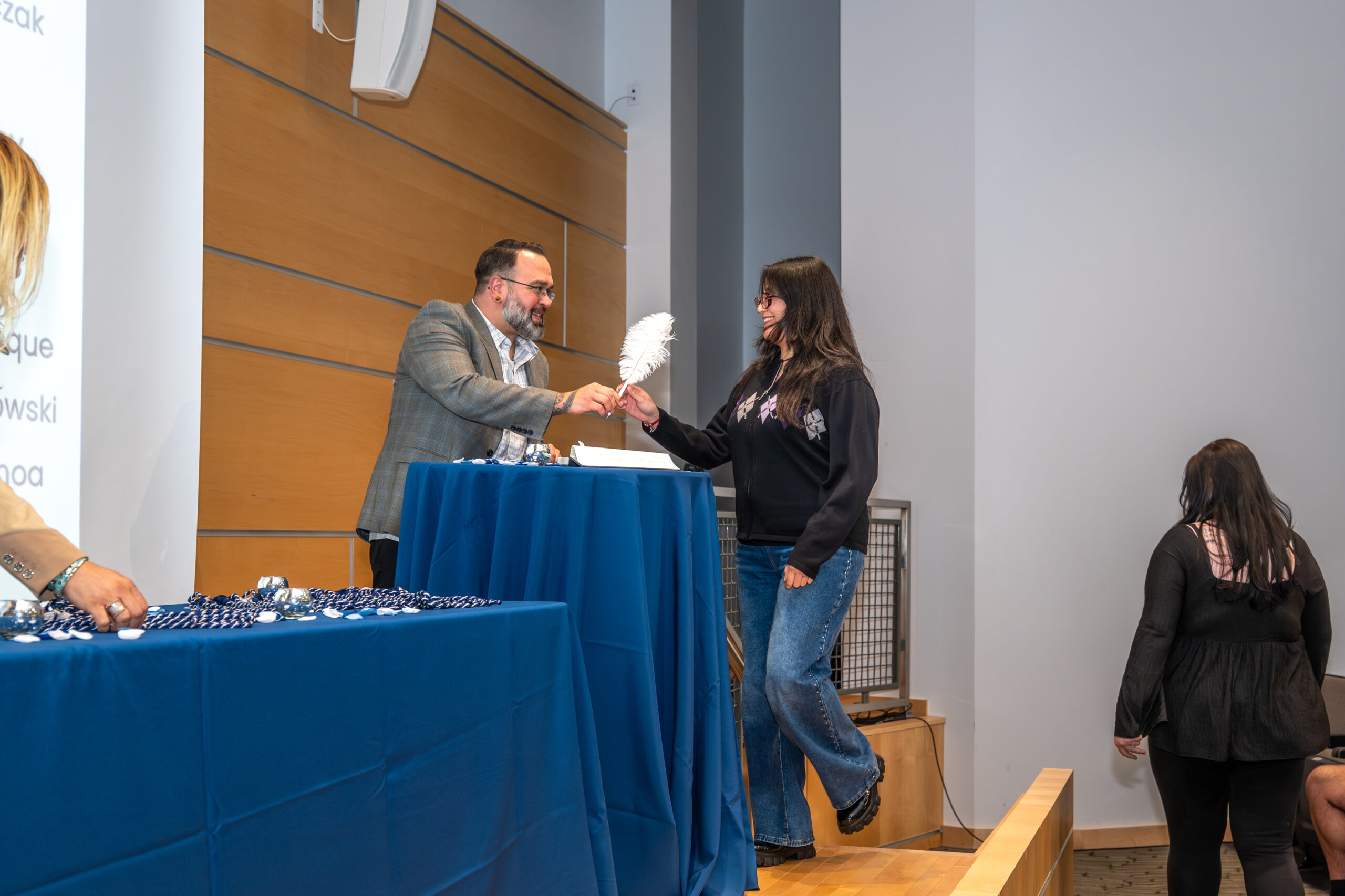 Man on stage handing a feather pen to a female student to sign an induction book