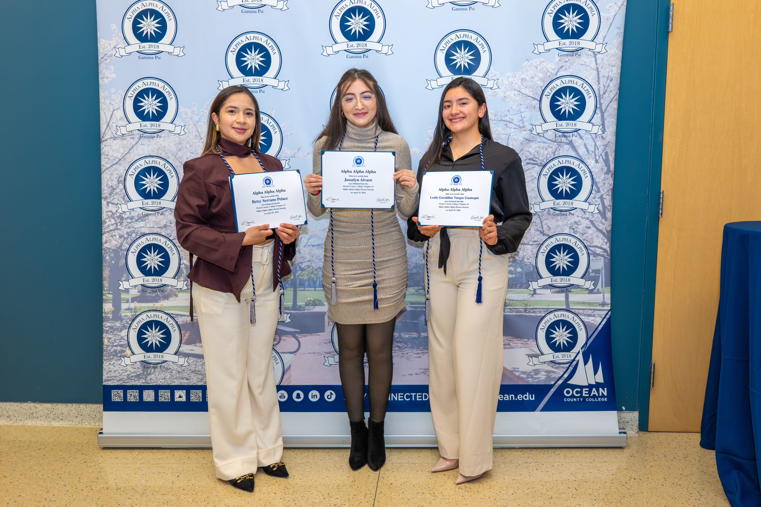 group of three female students posing in front of a step and repeat holding their honor society certificates.