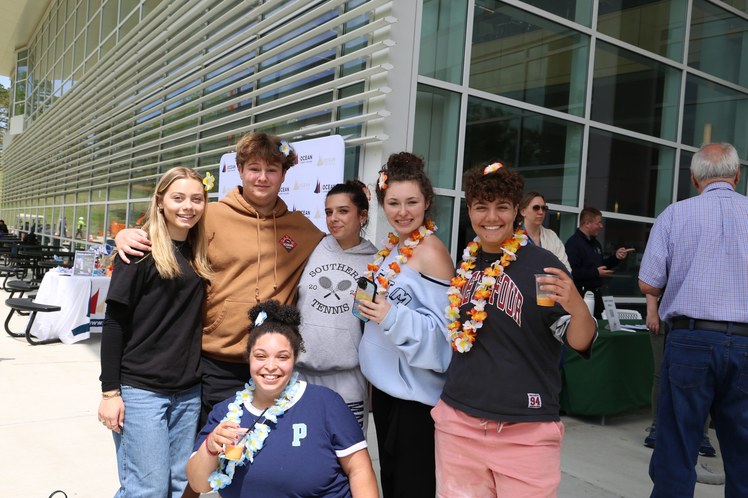 Group of smiling teens wearing leis posing outside a modern glass building at a student event.