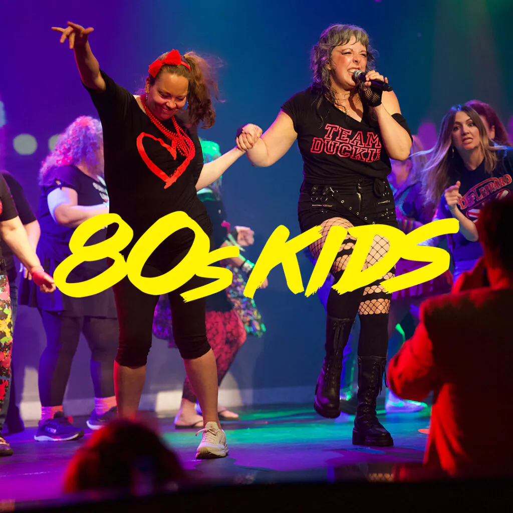 Two female performers on a brightly lit stage holding hands and dancing, with a large yellow '80s KIDS' logo across the foreground.