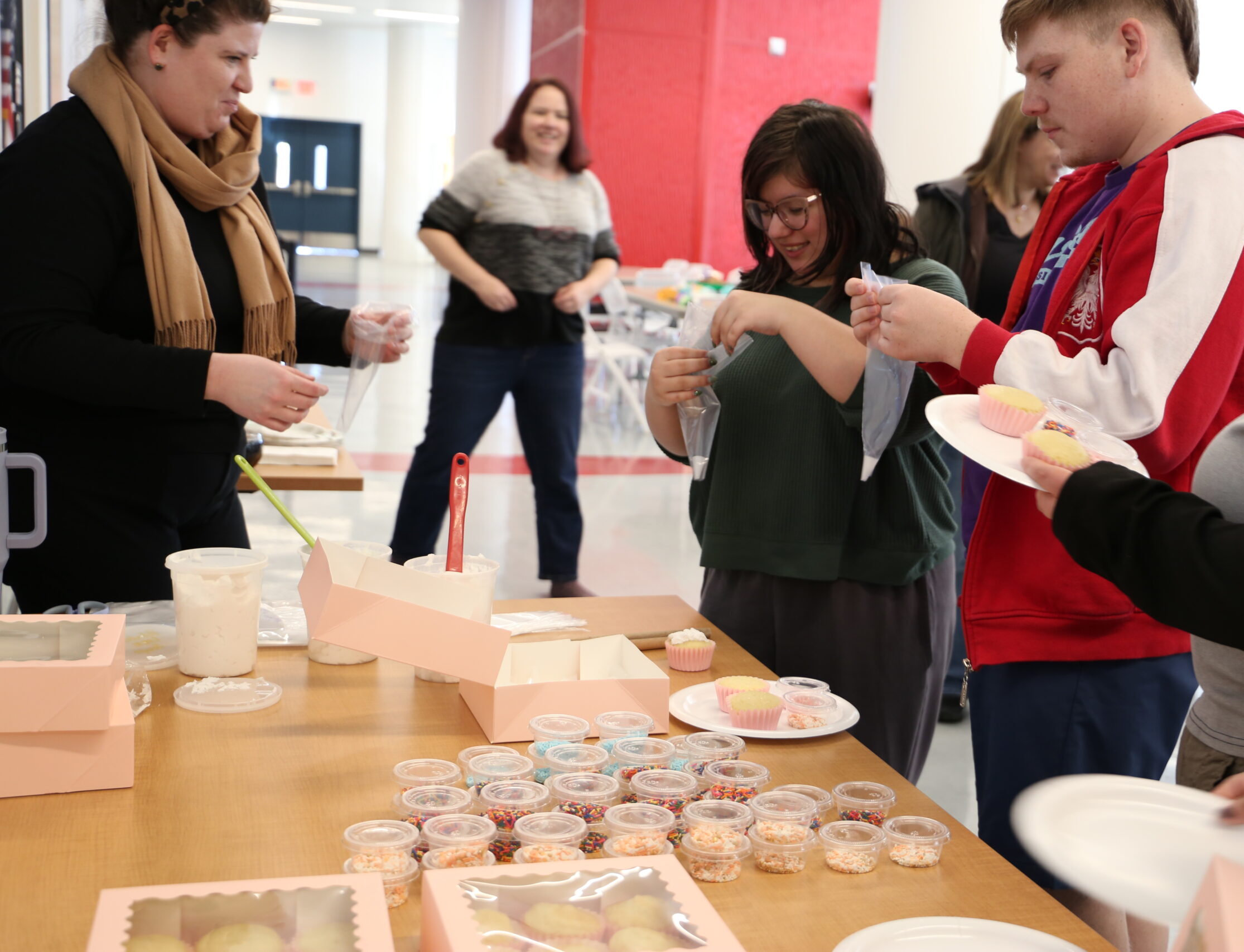 People gather around a table decorating cupcakes with sprinkles, pink boxes and frosting cups visible in the foreground.