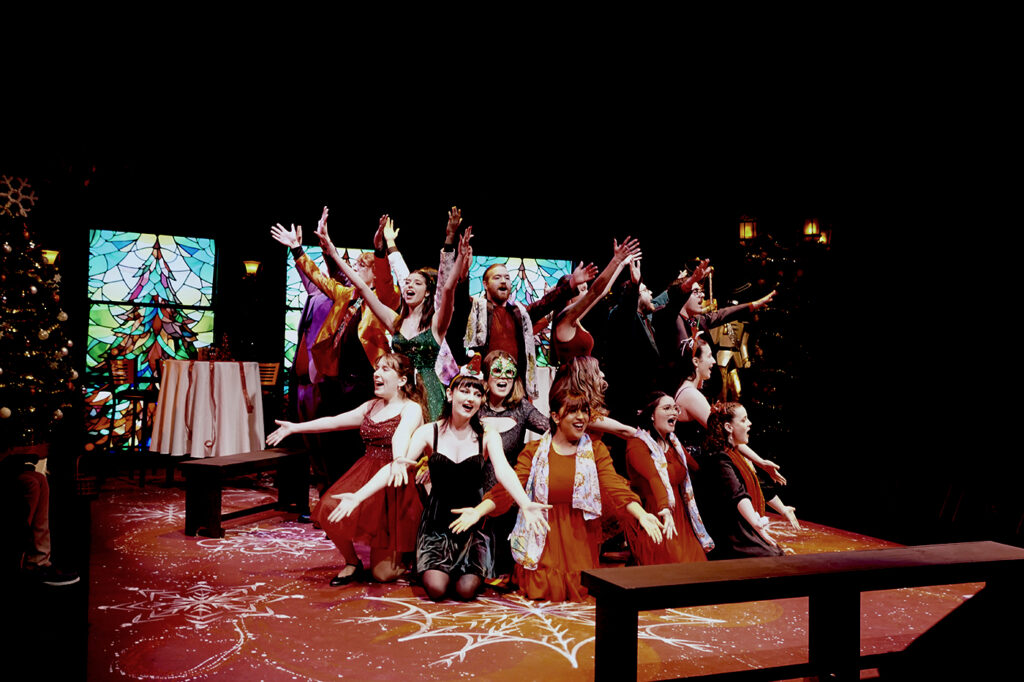 Group of performers in colorful costumes on a decorated stage, arms raised in a holiday musical number.