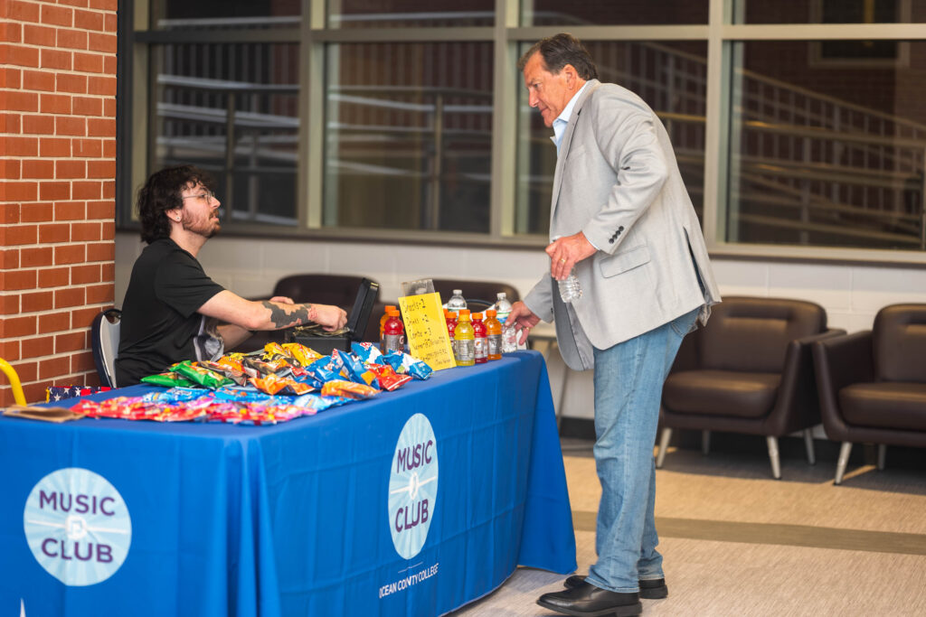 Man in a gray blazer hands a bottle of water to a seated volunteer at a snack table marked 'Music Club' inside a brick-walled venue.