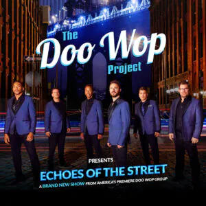 Six men in blue suits on a cobblestone street with streaks of light and surrounding buildings at night.