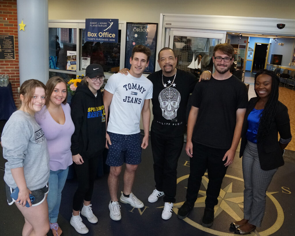 Group of seven young adults smiling and posing for a photo in a museum or science center lobby, with posters and a box office visible in the background.