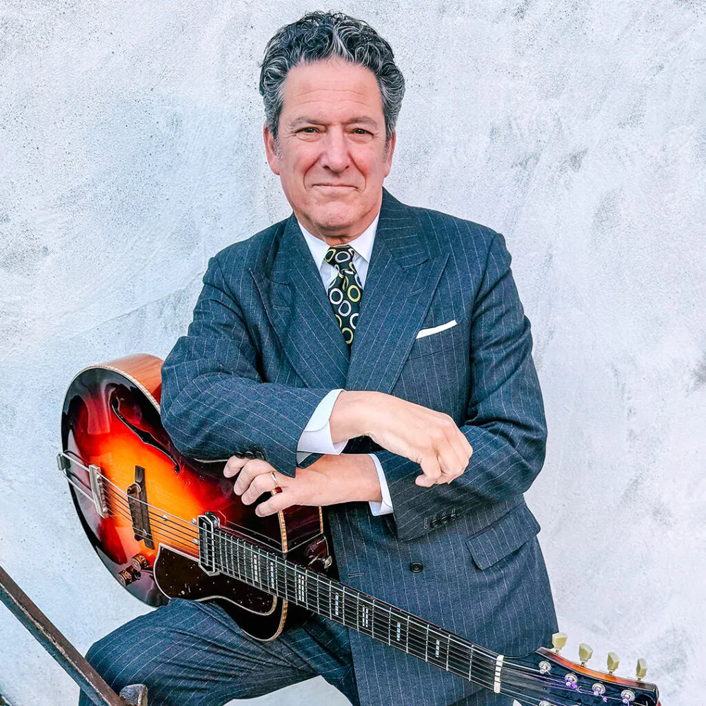 Man in a suit holding an acoustic guitar against a grey background.