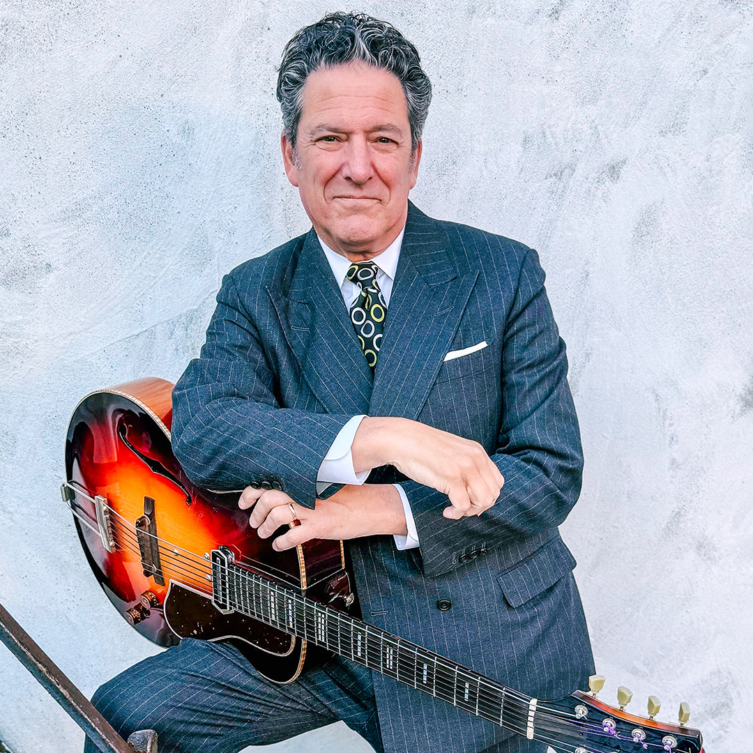 Man in a suit holding an acoustic guitar against a grey background.