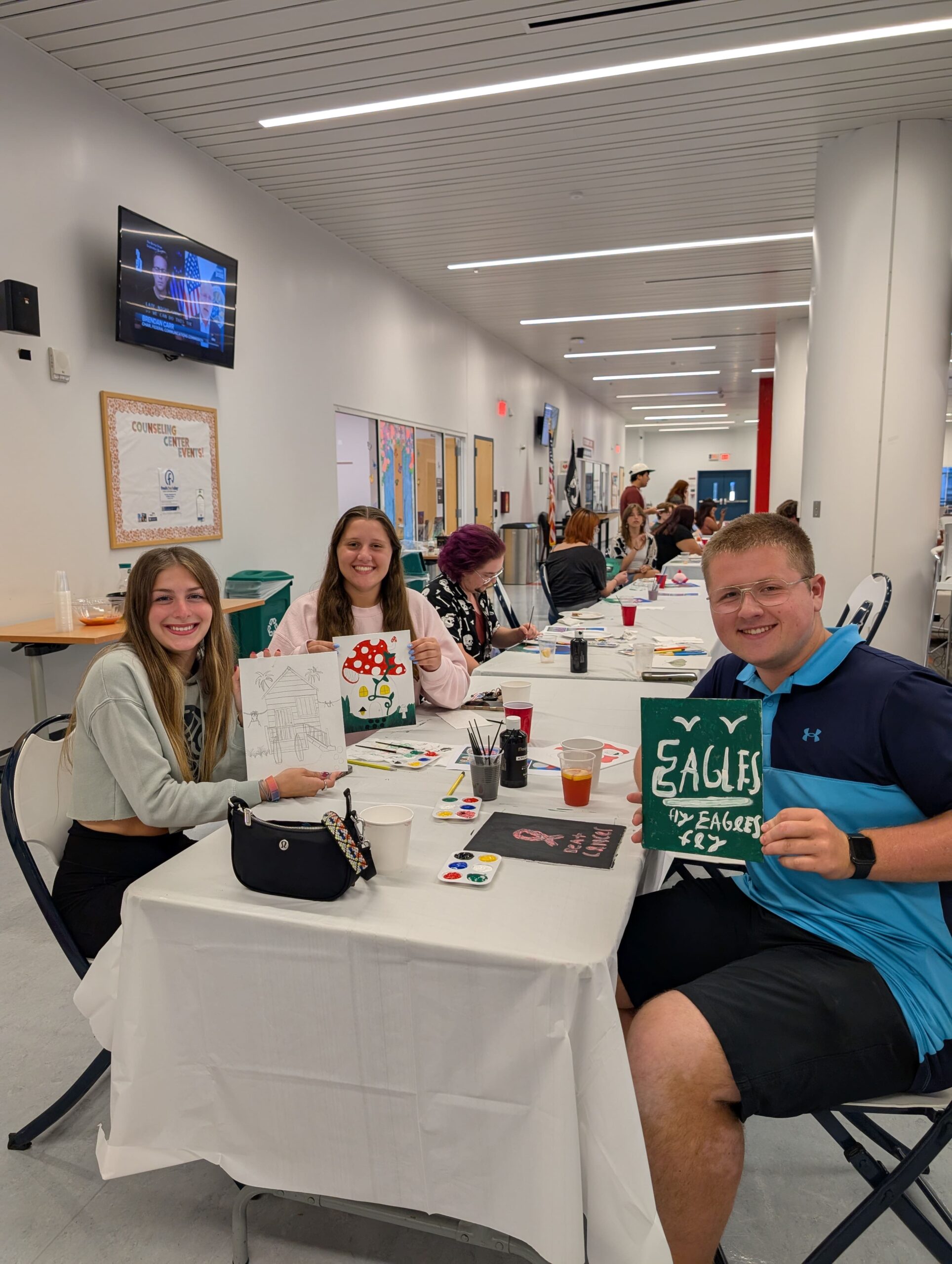 Smiling people sit at a long table painting and drawing at a craft event in a bright hallway; a man in a blue shirt holds a green sign that reads 'SALES EAGLES.'