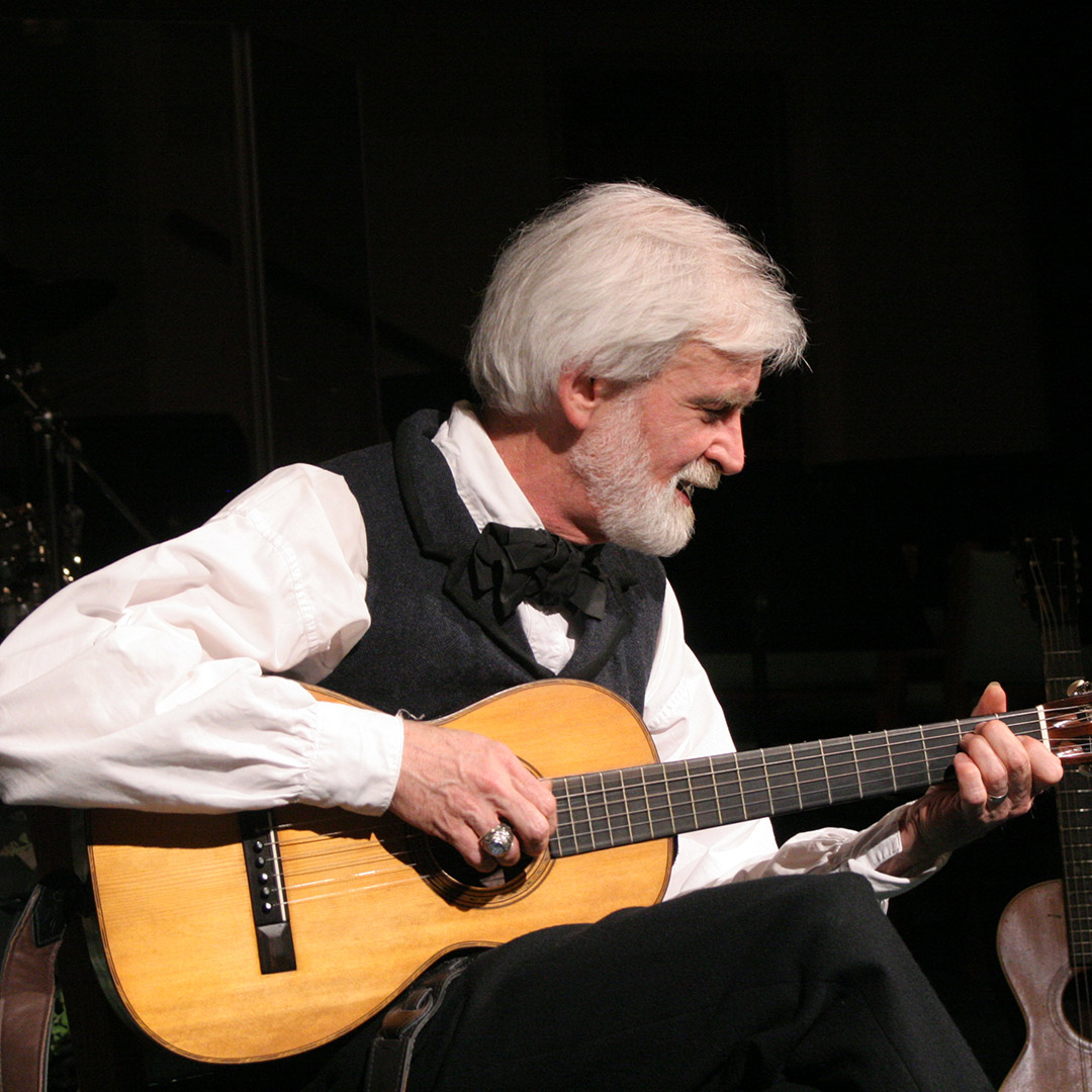 White-haired, bearded man in a vest and bow tie playing an acoustic guitar on a dark stage.