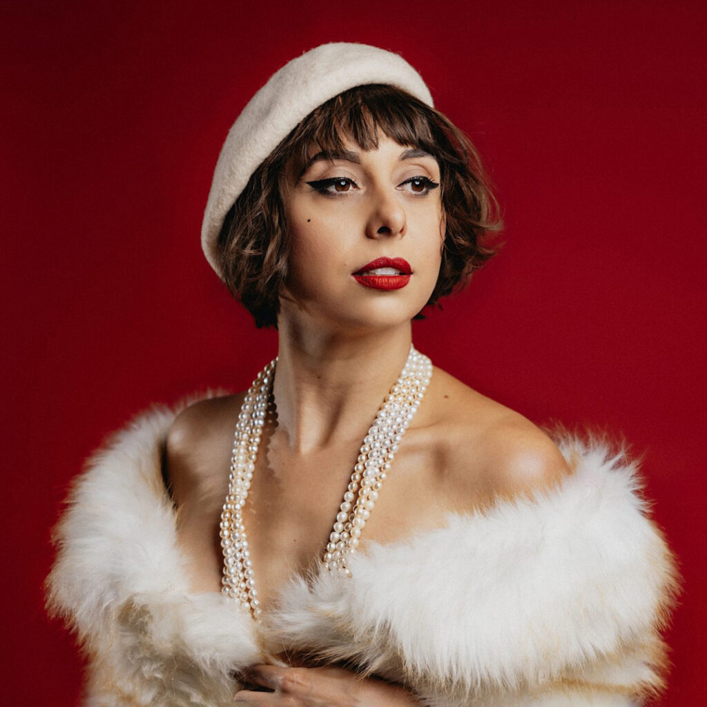 Portrait of a woman in a white beret, pearl necklaces, and white faux fur against a deep red background.
