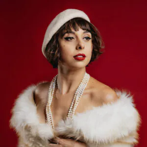 Portrait of a woman in a white beret, pearl necklaces, and white faux fur against a deep red background.