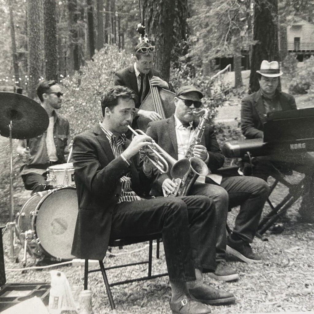 A jazz band performs outdoors in a forest, featuring a drummer, trumpet player, saxophonist, bassist, and keyboardist.